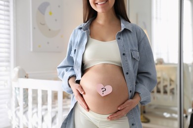 Pregnant woman with heart shaped sticky note on belly at home, closeup. Choosing baby name