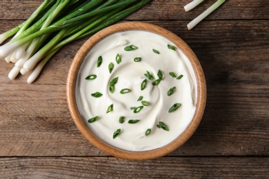 Bowl of fresh sour cream with green onion on wooden table, flat lay
