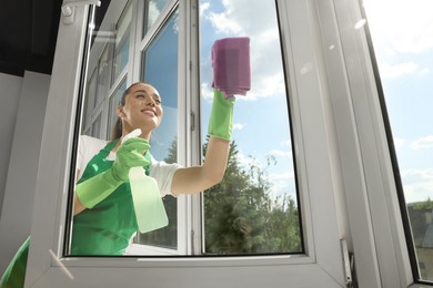 Happy young woman cleaning window glass with rag and spray indoors, low angle view