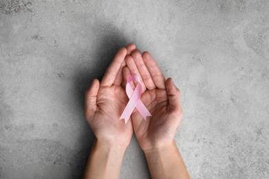 Woman holding pink ribbon at grey table, top view. Breast cancer awareness concept