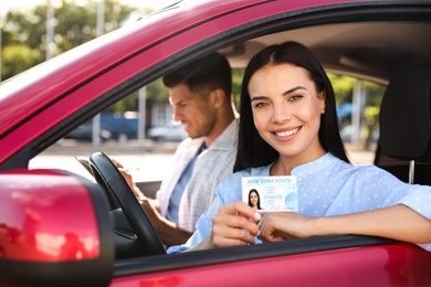 Photo of Young woman holding license while sitting in car with instructor. Driving school