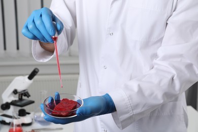 Scientist dripping red liquid into Petri dish with raw cultured meat in laboratory, closeup