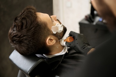 Professional hairdresser applying shaving foam onto client's skin in barbershop