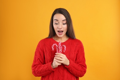Surprised young woman in red sweater holding candy canes on yellow background. Celebrating Christmas