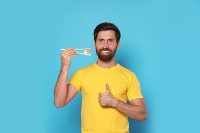 Happy man holding sushi roll with chopsticks and showing thumbs up on light blue background