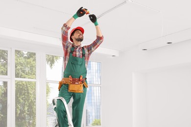 Photo of Electrician in uniform with pliers repairing ceiling wiring indoors