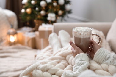 Woman with delicious hot drink wrapped in plaid on sofa at home, closeup. Space for text
