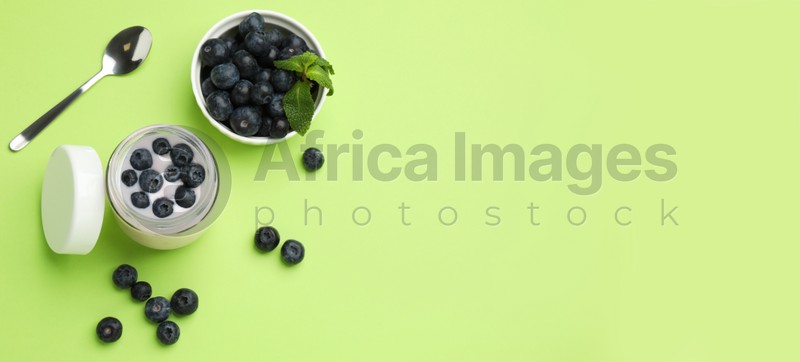 Jar of fresh yogurt with blueberries and spoon on light green background, flat lay with space for text. Banner design