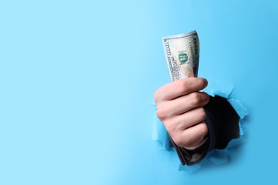 Businessman breaking through light blue paper with money in fist, closeup. Space for text