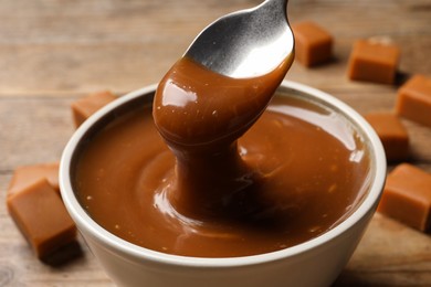 Photo of Taking yummy salted caramel with spoon from bowl at wooden table, closeup