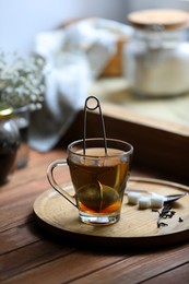 Photo of Cup of freshly brewed tea with snap infuser and sugar cubes on wooden table indoors