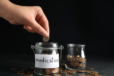 Woman putting coin into glass jar with tag MEDICATION on black table, closeup