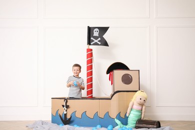 Cute little boy playing in pirate cardboard ship near white wall indoors