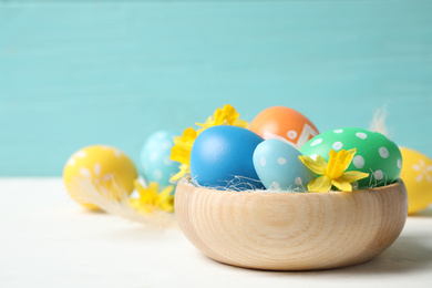Colorful Easter eggs and flowers in bowl on white wooden table. Space for text