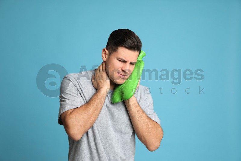 Man using hot water bottle to relieve neck pain on light blue background