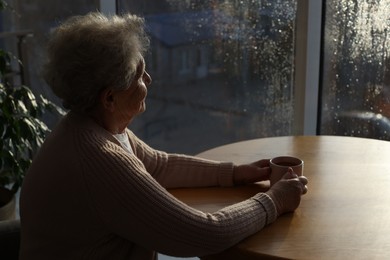 Elderly woman with drink looking out of window indoors on rainy day, space for text. Loneliness concept
