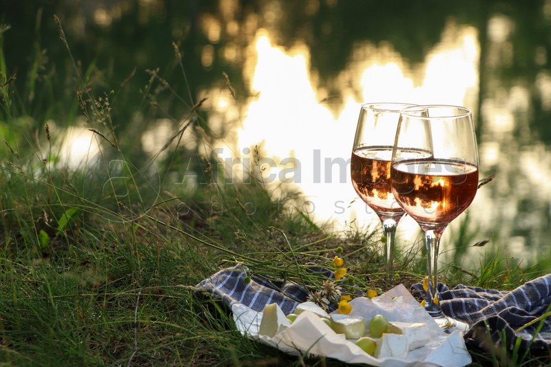 Glasses of delicious rose wine, cheese and grapes on picnic blanket near lake, space for text