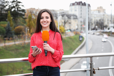 Young female journalist with microphone and smartphone working on city street. Space for text