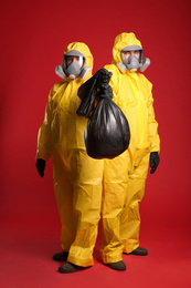 Man and woman in chemical protective suits holding trash bag on red background. Virus research