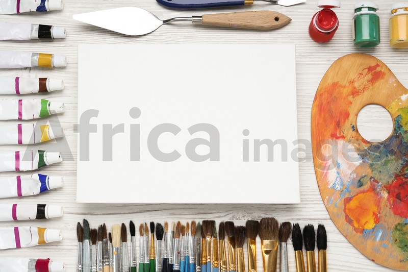 Blank canvas, brushes, palette and paints on white wooden table, flat lay. Space for text
