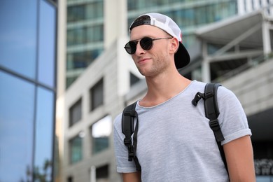 Handsome young man with stylish sunglasses and backpack near building outdoors, space for text
