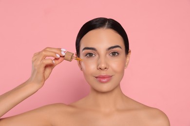 Young woman applying essential oil onto face against pink background