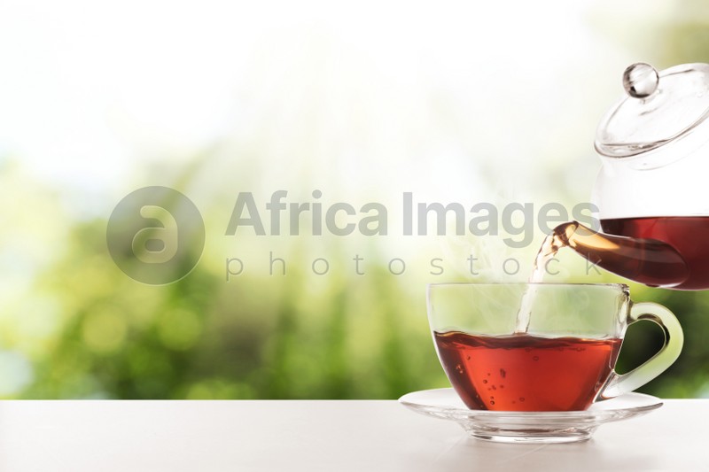 Pouring fresh hot tea into glass cup on table against blurred green background. Space for text