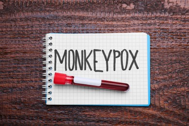 Word Monkeypox written in notebook and test tube with blood sample on wooden table, top view