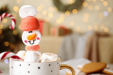 Funny marshmallow snowman in cup of hot drink against blurred festive lights, closeup. Space for text
