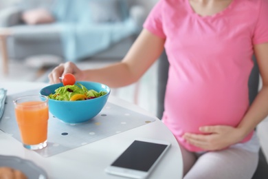 Young pregnant woman eating vegetable salad at table in kitchen