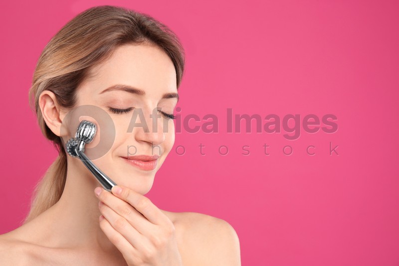 Photo of Young woman using metal face roller on pink background, space for text