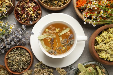 Cup of aromatic freshly brewed tea surrounded by different dry herbs on grey table, flat lay