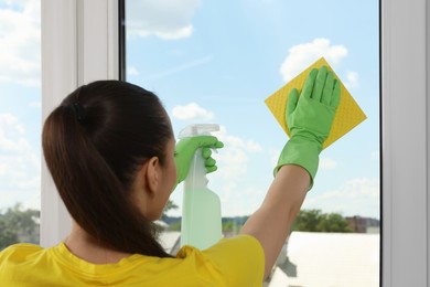 Woman cleaning window glass with sponge cloth and spray indoors