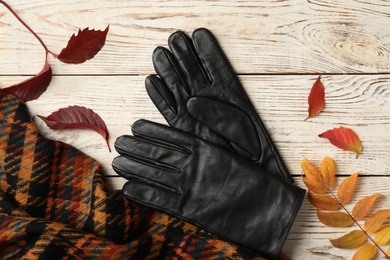 Stylish black leather gloves, scarf and dry leaves on white wooden table, flat lay