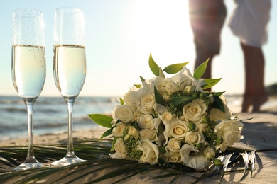 Beautiful wedding bouquet, glasses of champagne and couple on sandy beach