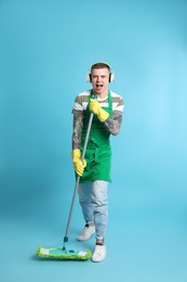 Handsome young man with headphones and mop singing on light blue background