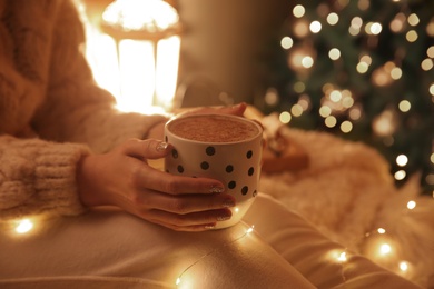 Woman with cup of hot drink near Christmas tree at home, closeup