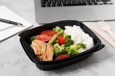 Photo of Container with tasty food, laptop, cutlery and notebook on light grey table. Business lunch