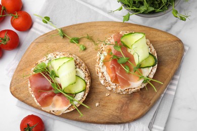 Crunchy buckwheat cakes with cream cheese, prosciutto and cucumber slices on white marble table, flat lay