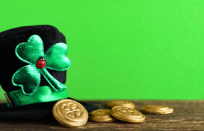 Black leprechaun hat and gold coins on wooden table against green background, space for text. St. Patrick's Day celebration