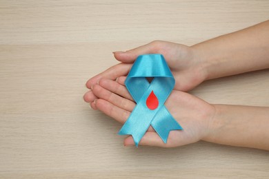 Photo of Woman holding light blue ribbon with paper blood drop at wooden table, top view. Diabetes awareness