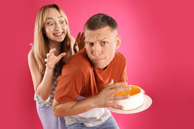 Greedy man hiding tasty cake from woman on pink background