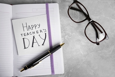 Paper with inscription HAPPY TEACHER'S DAY, notebook and eyeglasses on grey table, flat lay