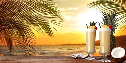 Tasty Pina Colada cocktail on wooden table near ocean at sunset, space for text. Banner design