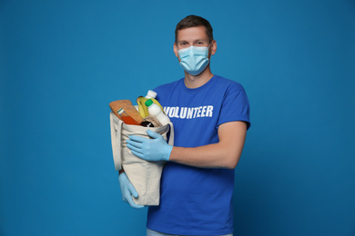 Male volunteer in protective mask and gloves with products on blue background. Aid during coronavirus quarantine	