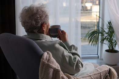 Elderly woman with cup of drink sitting in armchair indoors, space for text. Loneliness concept