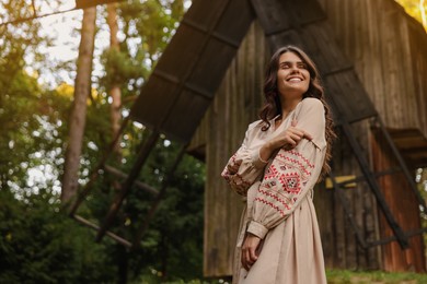 Beautiful woman wearing embroidered dress near old wooden mill in countryside. Ukrainian national clothes