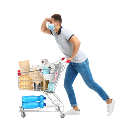 Young man in medical mask and shopping cart with purchases on white background. Coronavirus pandemic 