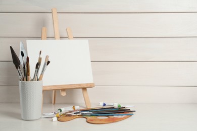 Easel with blank canvas, brushes, paints and palette on white wooden table. Space for text