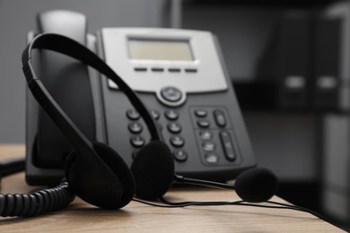 Modern headset near stationary phone on wooden desk in office, closeup. Hotline service